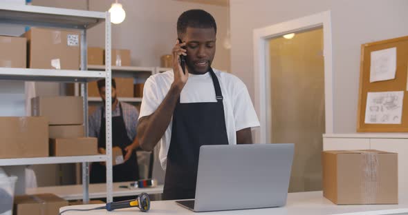 African Man Talking on Phone Using Laptop Working at Warehouse for Online Seller