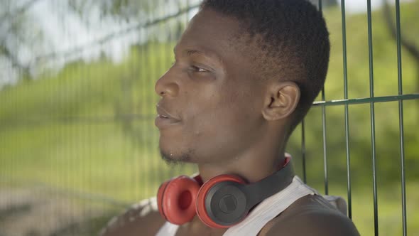 Close-up of African American Sportsman Sitting at Mesh Fence and Smiling. Side View Portrait of alt