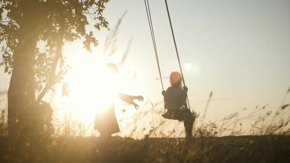 Silhouette of Happy Young Mother and Little Daughter on a Swing at Sun Light