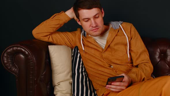 Young Man Browsing Mobile Phone Sitting on Sofa Leaning on Pillows