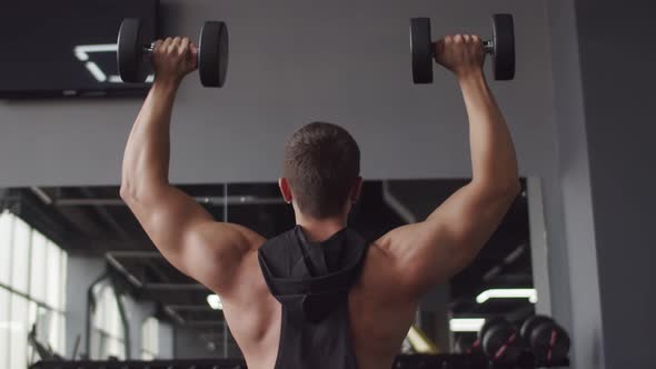 Young male bodybuilder trains in gym, lifting two dumbbells with two hands, back view. Male athlete