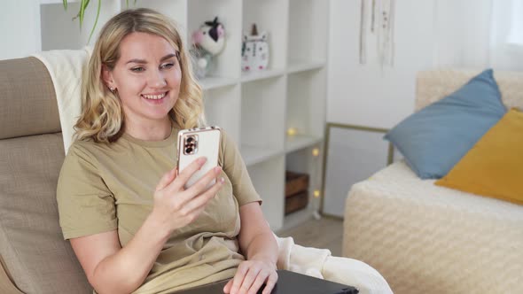 Cute young blonde woman uses phone while sitting at home in armchair Using technology concept