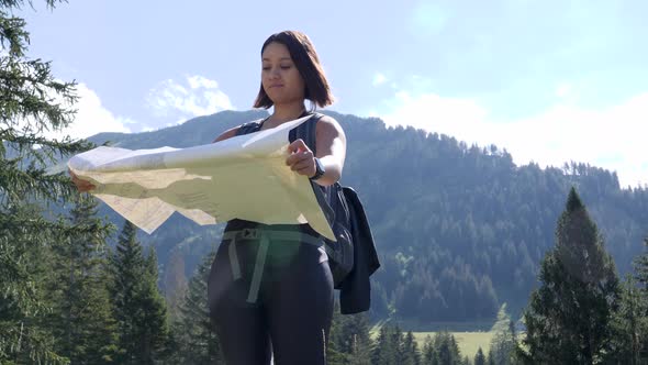 Young female hiker with backpack spreads foldable map in sunny mountain landscape and looks for the