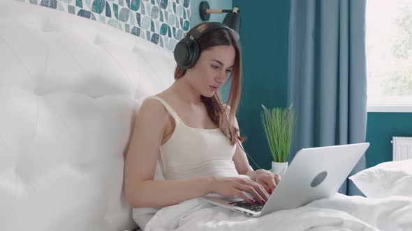 Brunette Woman in Headphone Typing Message in Computer on Bed