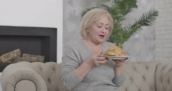 Portrait of Happy Senior Caucasian Woman with Brown Eyes Smelling and Biting Delicious Croissant