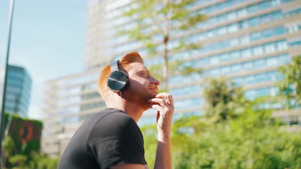 Pensive red haired man listening music in headphones while sitting on the bench