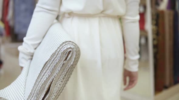 Fashionable Young Woman in White Clothes Walking Through the Textile Store with Fabric Roll