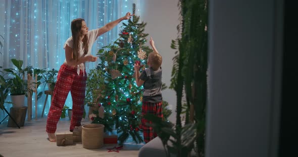 A Young Mother and Two Boys in Pajamas Decorate the Christmas Tree with Toys on New Year's Eve