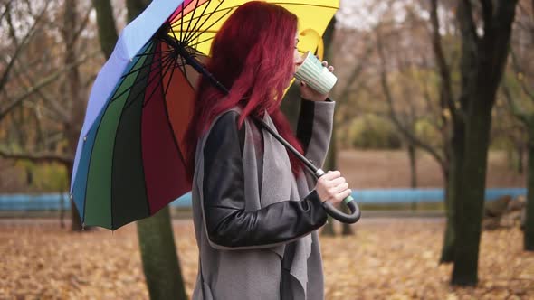 Side View Young Woman with Red Hair Walking in Autumn Park and Drinking Coffee From a Paper Cup