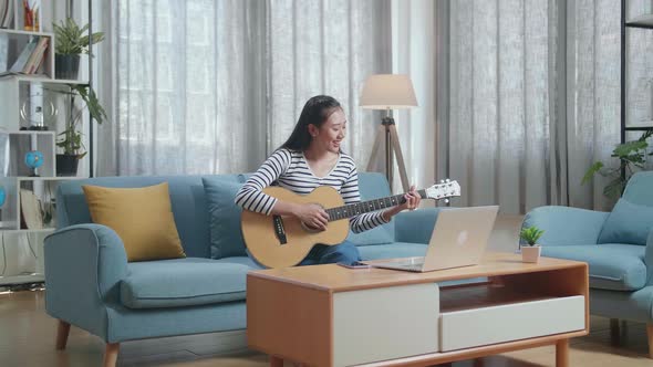 Smiling Asian Woman Enjoy Learning Playing Guitar By A Laptop At Home