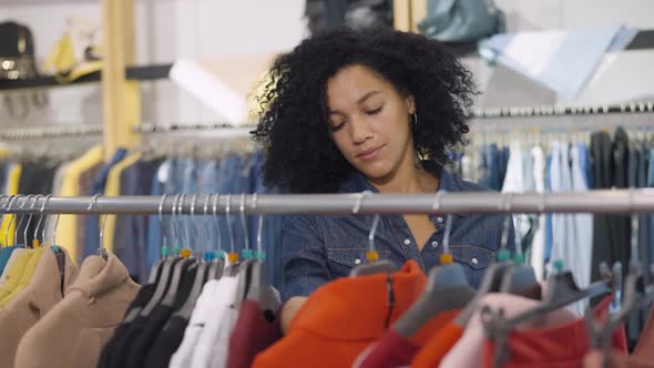 Female Customer Makes Purchases in Clothing Store Chooses Stylish Clothes Hanging on Rack Hangers