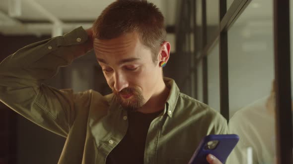 A Handsome Young Student Stands Near the Window Keeps a Phone in His Hands and Reads Something