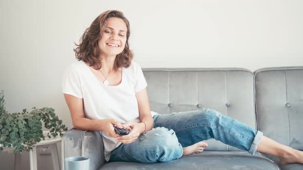 Young Woman Sitting on a Sofa with a TV Remote Control and Switching Channels