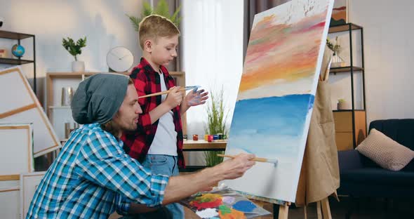 Male Artist Teaching Small Boy to Draw Picture in Home Studio Using Canvas on Easel