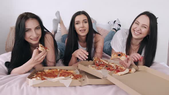 Three Gorgeous Girlfriends Are Lying on the Bed and Eating Delivery Pizza