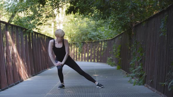 Middleaged Blonde Caucasian Woman Athlete Lady Wears Black Tracksuit Stands on Bridge in City Park