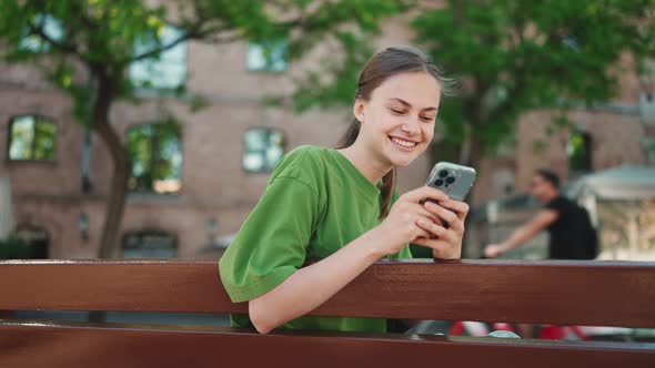 Pleased blonde woman wearing green t-shirt texting by phone on the bench