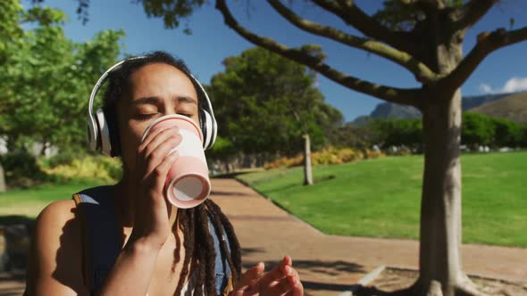 African american woman wearing headphones sitting drinking coffee in park