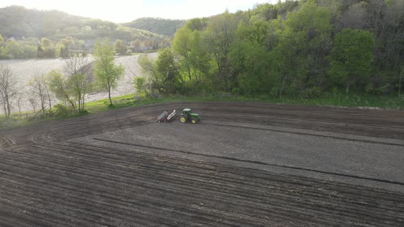 Farmer driving tractor fertilizing and seeding section of farmland in valley with cloudy sky.