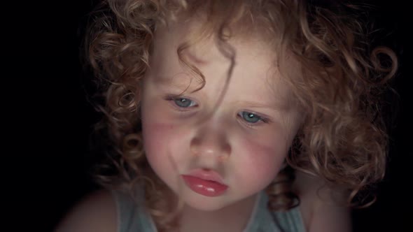 Close up face of a little caucasian curly girl looking at the screen of a digital device at night.