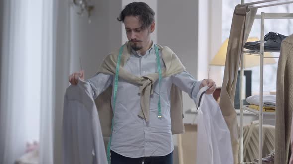 Portrait of Puzzled Young Handsome Caucasian Man Comparing Shirts on Hangers Thinking