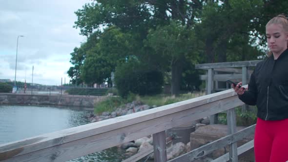 Slow motion. Girl in sportswear walking on wooden bridge and taking pictures with her smartphone.