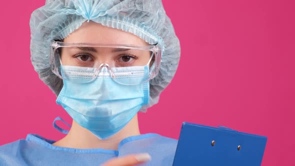 Serious Doctor in a Blue Medical Coat Safety Glasses and Medical Cap Looking at Camera