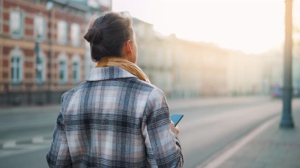 Back View of a Businesswoman in a Coat Walking Around the Old City at the Dawn and Using Smartphone