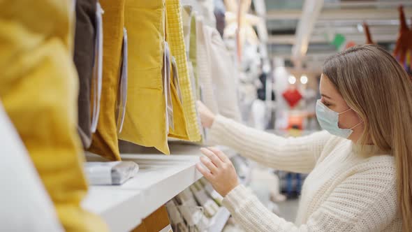 Woman in a Protective Mask Chooses Decorative Pillows in a Store with Household