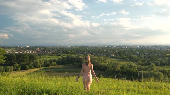 Young woman in pink dress walking on green summer field outdoors.