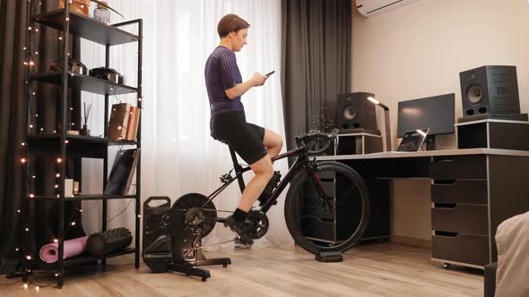 Woman is cycling on exercise bicycle with smartphone in hands.