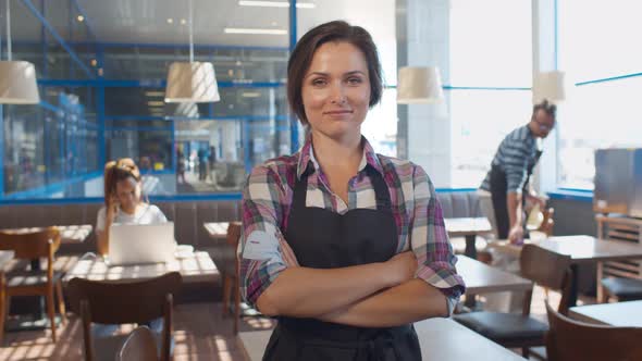 Portrait of Cheerful Waitress in Apron Smiling at Camera with Hands Crossed Standing in Cafe