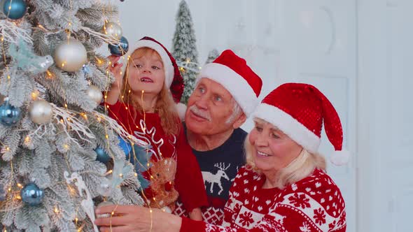 Toddler Girl with Senior Couple Grandparents Decorating Artificial Christmas Pine Tree at Home Room
