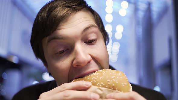 A Man with Appetite Bites a Hamburger on a Food Court in a Shopping Center Closeup