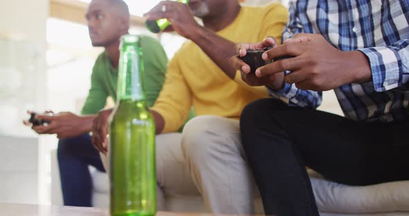 African american father and twin teenage sons sitting on couch playing game on tv and drinking beer