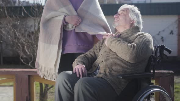 Positive Old Caucasian Disabled Man in Wheelchair Sitting Outdoors in Sunrays and Talking with