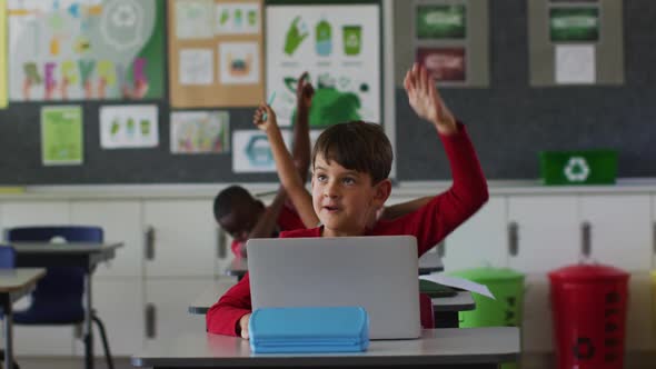 Portrait of happy caucasian schoolboy with laptop sitting raising hand to answer questions