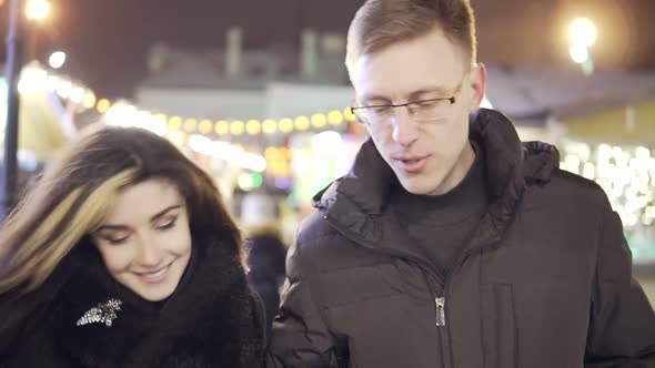 Young Couple Walk Togerher Near Christmas Lights at Christmas Market with a Cup of Warm Tea