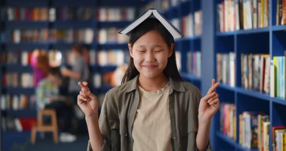 Superstitious Asian Kid Holding School Book on Head Keeping Fingers Crossed in Library