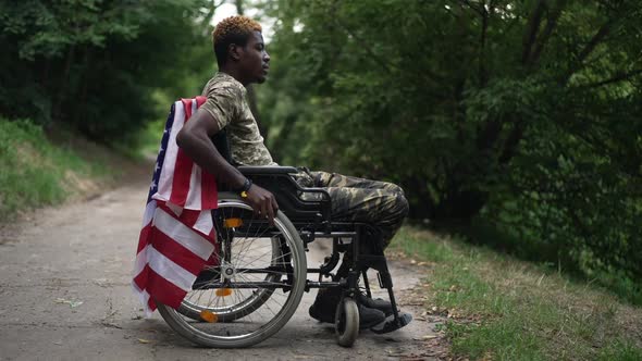 Side View Thoughtful Depressed African American Young Soldier in Wheelchair with USA Flag Looking