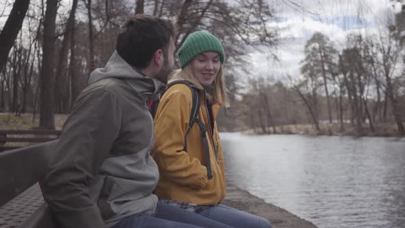 A Young Couple of Tourists with Backpacks Sitting on the Bench in the Spring Park at the Riverbank