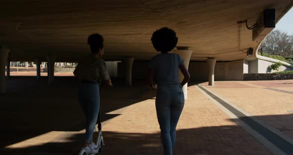 Two mixed race women riding electric scooter under bridge