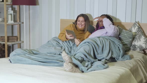 African-American and European woman Lying in Bed Using a Tablet At Home, Evening, Smiling