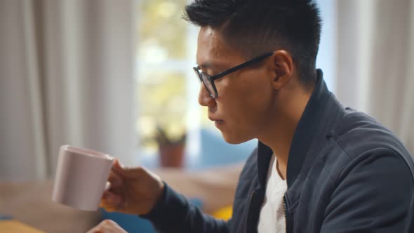 Close Up of Young Asian Man Working on Laptop Drinking Coffee at Home Office