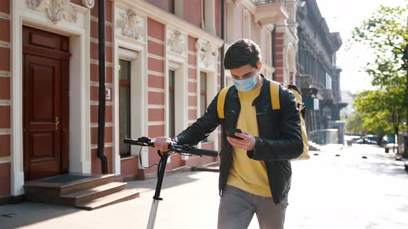 Delivery Man with Yellow Bagpack and Protective Mask Walking with Electric Scooter with Food