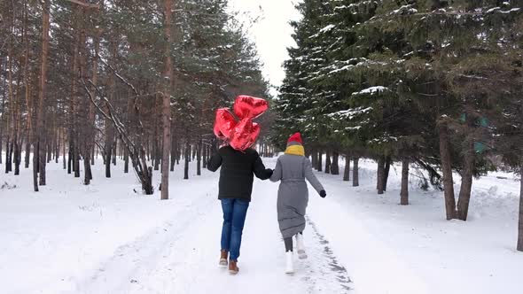 Young Couple Holding Hands Together While Running Away with Red Heart Shape Balloons