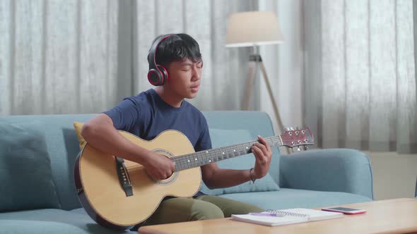 Asian Boy Composer With Notebook On Table Wearing Headphones Singing And Playing Guitar At Home