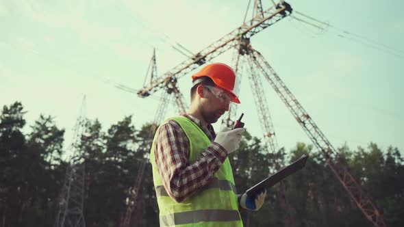 Electrical Equipment Worker Near High Voltage Tower Using Walkie Talkie and Clipboard