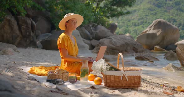 Young Woman in Hat and Yellow Dress Working on Laptop Sitting on Beach Perfect Summer Day Outdoors