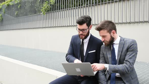 Two young bearded men sitting outdoor discussing using personal computer